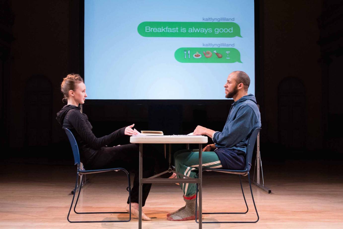 Kaitlyn Gilliland and Will Rawls sitting at a table in #loveyoumeanit with a projection of their text messages in the background.